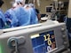 A group of medical professionals wearing blue surgical attire performing a surgery. In the foreground, there is a medical monitor displaying vital signs. The background includes medical equipment like IV drips, monitors, and respiratory devices. The setting appears to be a hospital operating room.