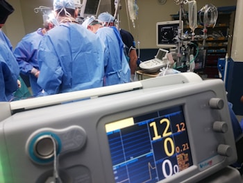 A group of medical professionals wearing blue surgical attire performing a surgery. In the foreground, there is a medical monitor displaying vital signs. The background includes medical equipment like IV drips, monitors, and respiratory devices. The setting appears to be a hospital operating room.