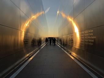 A pathway flanked by tall reflective walls with engraved names. The golden hue of the setting sun casts a soft glow, creating an arch-like reflection on the walls. A small group of people stands at the end of the path, silhouetted by the light.