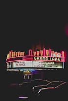 Neon lights illuminate the marquee of a classic theater, displaying movie titles like 'The Force' and 'Logan Lucky'. Bright red and yellow colors create a nostalgic atmosphere against the dark night sky, with a glimpse of parked cars below.