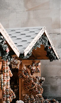 A festive market stall featuring a sloped roof adorned with green garlands and small red ornaments. The stall is decorated with an assortment of dried fruit and nut wreaths, shaped into stars and other designs.
