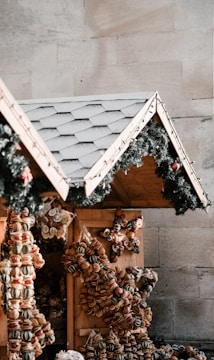A festive market stall featuring a sloped roof adorned with green garlands and small red ornaments. The stall is decorated with an assortment of dried fruit and nut wreaths, shaped into stars and other designs.
