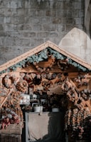 A cozy wooden market stall displays an array of decorative wreaths and ornaments made from dried fruits and other natural materials. Pine branches with pine cones adorn the top of the stall, adding a festive touch. The stall is set against a textured stone wall, offering a rustic and charming atmosphere.