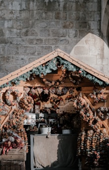 A cozy wooden market stall displays an array of decorative wreaths and ornaments made from dried fruits and other natural materials. Pine branches with pine cones adorn the top of the stall, adding a festive touch. The stall is set against a textured stone wall, offering a rustic and charming atmosphere.