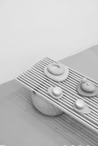 A grayscale artistic arrangement featuring smooth, rounded stones placed on a slatted wooden surface. The surface angles slightly, creating dynamic shadows, while a larger round stone supports the entire structure. The minimalistic composition highlights textures and shapes, conveying a sense of calm balance.