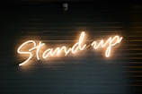 A neon light sign displaying the words 'Stand up' in cursive script against a dark, horizontal slatted background.