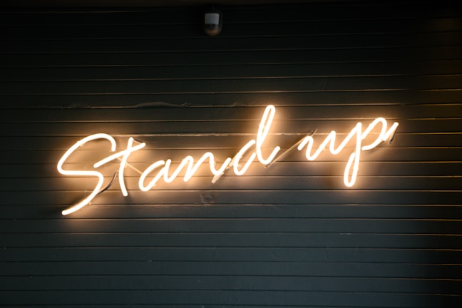 A neon light sign displaying the words 'Stand up' in cursive script against a dark, horizontal slatted background.
