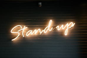 A neon light sign displaying the words 'Stand up' in cursive script against a dark, horizontal slatted background.