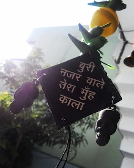 A decorative hanging with a black plaque containing text in Hindi, possibly meant as a protective charm against evil. It is adorned with small skull-shaped ornaments and green peppers, and there is a lemon above. The background shows blurred greenery and a railed staircase, with bright sunlight creating a high contrast.