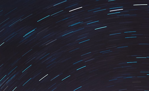 Long exposure photograph of the night sky capturing star trails in an arc pattern. These trails create a dynamic sense of movement against a dark backdrop.
