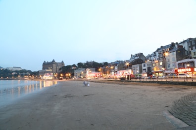 A seaside townscape featuring a sandy beach in the foreground with a calm sea to the left. Along the right, a row of illuminated buildings and a grand hotel create a lively, bustling atmosphere. Streetlights cast a warm glow over the scene as dusk approaches, and a few people are visible walking on the beach.