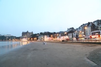 A seaside townscape featuring a sandy beach in the foreground with a calm sea to the left. Along the right, a row of illuminated buildings and a grand hotel create a lively, bustling atmosphere. Streetlights cast a warm glow over the scene as dusk approaches, and a few people are visible walking on the beach.
