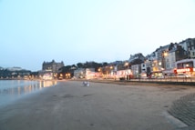 A seaside townscape featuring a sandy beach in the foreground with a calm sea to the left. Along the right, a row of illuminated buildings and a grand hotel create a lively, bustling atmosphere. Streetlights cast a warm glow over the scene as dusk approaches, and a few people are visible walking on the beach.