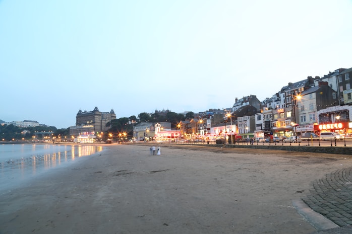 A seaside townscape featuring a sandy beach in the foreground with a calm sea to the left. Along the right, a row of illuminated buildings and a grand hotel create a lively, bustling atmosphere. Streetlights cast a warm glow over the scene as dusk approaches, and a few people are visible walking on the beach.