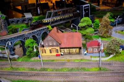 A miniature model railway scene featuring a detailed model train crossing a stone viaduct. Below, there are small model buildings, including a station with intricate architectural features and trees surrounding the area. The tracks are meticulously arranged through the green landscape.