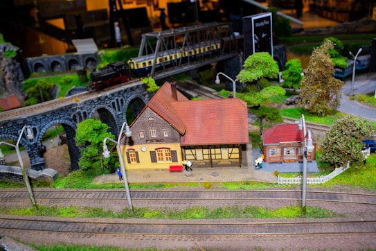 A miniature model railway scene featuring a detailed model train crossing a stone viaduct. Below, there are small model buildings, including a station with intricate architectural features and trees surrounding the area. The tracks are meticulously arranged through the green landscape.