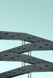 A modern architectural structure featuring repetitive patterns of horizontal concrete beams with small rectangular openings, supported by vertical metal beams against a clear, pale blue sky.