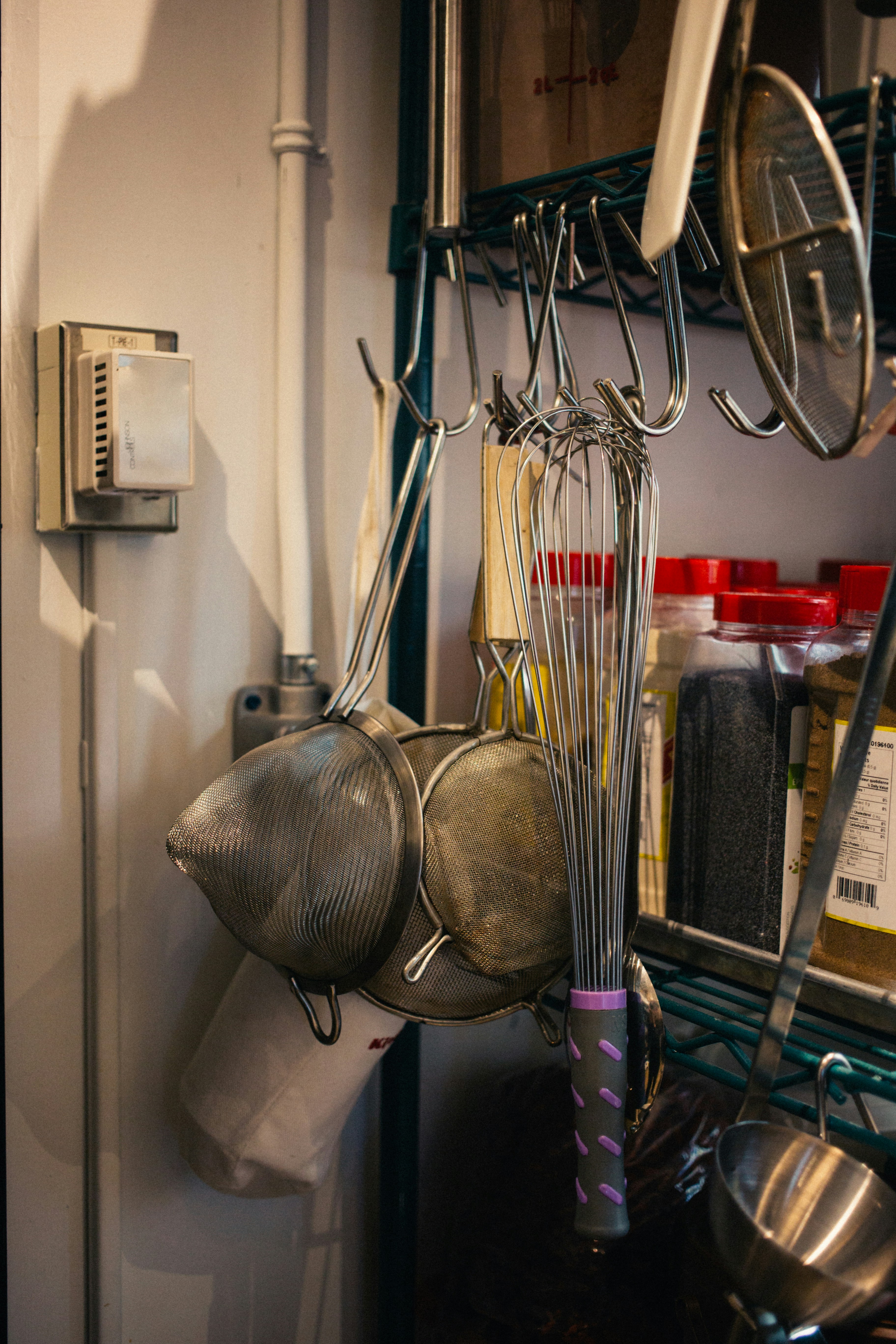 A kitchen setup with metal whisks and strainers hanging on hooks. There are jars with red lids containing spices or ingredients on a shelf. The background shows a metal rack and a thermostat on the wall.