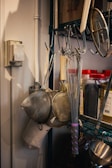 A kitchen setup with metal whisks and strainers hanging on hooks. There are jars with red lids containing spices or ingredients on a shelf. The background shows a metal rack and a thermostat on the wall.