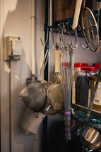 A kitchen setup with metal whisks and strainers hanging on hooks. There are jars with red lids containing spices or ingredients on a shelf. The background shows a metal rack and a thermostat on the wall.