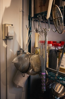 A kitchen setup with metal whisks and strainers hanging on hooks. There are jars with red lids containing spices or ingredients on a shelf. The background shows a metal rack and a thermostat on the wall.