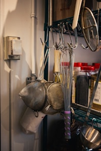 A kitchen setup with metal whisks and strainers hanging on hooks. There are jars with red lids containing spices or ingredients on a shelf. The background shows a metal rack and a thermostat on the wall.