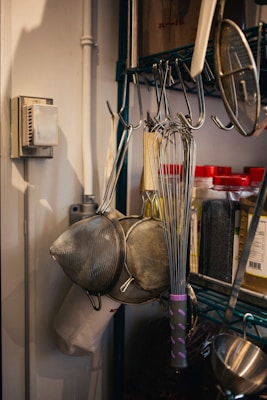 A kitchen setup with metal whisks and strainers hanging on hooks. There are jars with red lids containing spices or ingredients on a shelf. The background shows a metal rack and a thermostat on the wall.