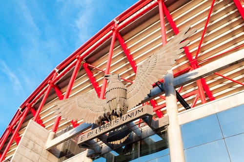 A large eagle sculpture with outstretched wings is mounted above an entrance, set against a backdrop of red structural beams and a modern building facade. The phrase 'E Pluribus Unum' is displayed below the eagle.