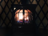 A cozy, lit fireplace with visible flames and logs inside. The fireplace is enclosed in a dark metal frame with arched glass panes. The background features a patterned wall with a crisscross design.