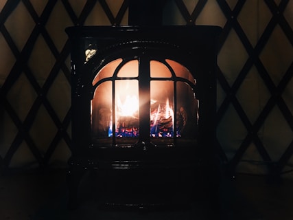 A cozy, lit fireplace with visible flames and logs inside. The fireplace is enclosed in a dark metal frame with arched glass panes. The background features a patterned wall with a crisscross design.
