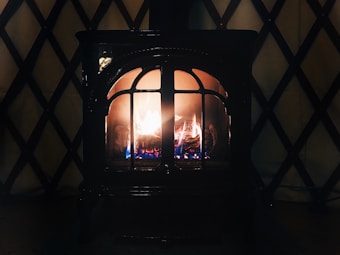 A cozy, lit fireplace with visible flames and logs inside. The fireplace is enclosed in a dark metal frame with arched glass panes. The background features a patterned wall with a crisscross design.