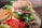 A delicious-looking grilled chicken burger topped with melted cheese, lettuce, tomato slices, cucumber, and sauce, served on a sesame seed bun. The burger is accompanied by a side of crispy golden French fries. Additional garnishes include slices of fresh tomato, black olives, sweet corn, and mushrooms on the side.