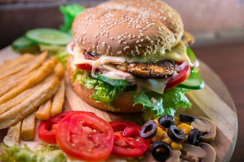 A delicious-looking grilled chicken burger topped with melted cheese, lettuce, tomato slices, cucumber, and sauce, served on a sesame seed bun. The burger is accompanied by a side of crispy golden French fries. Additional garnishes include slices of fresh tomato, black olives, sweet corn, and mushrooms on the side.