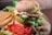A delicious-looking grilled chicken burger topped with melted cheese, lettuce, tomato slices, cucumber, and sauce, served on a sesame seed bun. The burger is accompanied by a side of crispy golden French fries. Additional garnishes include slices of fresh tomato, black olives, sweet corn, and mushrooms on the side.