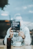 A person seated at a table is holding a book titled 'Faith Forward Future' in front of their face, with only their hands visible. They are wearing a light-colored hoodie and beaded bracelets. On the table, there is a tall glass of soda and a cup of coffee.