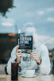 A person seated at a table is holding a book titled 'Faith Forward Future' in front of their face, with only their hands visible. They are wearing a light-colored hoodie and beaded bracelets. On the table, there is a tall glass of soda and a cup of coffee.
