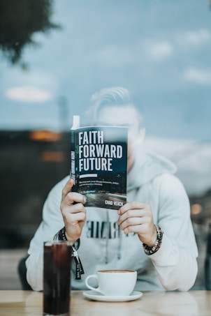 A person seated at a table is holding a book titled 'Faith Forward Future' in front of their face, with only their hands visible. They are wearing a light-colored hoodie and beaded bracelets. On the table, there is a tall glass of soda and a cup of coffee.