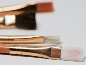 A close-up of three artist paintbrushes with golden ferrules laying on a light surface. The bristles vary in color and thickness, highlighting each brush's unique purpose.