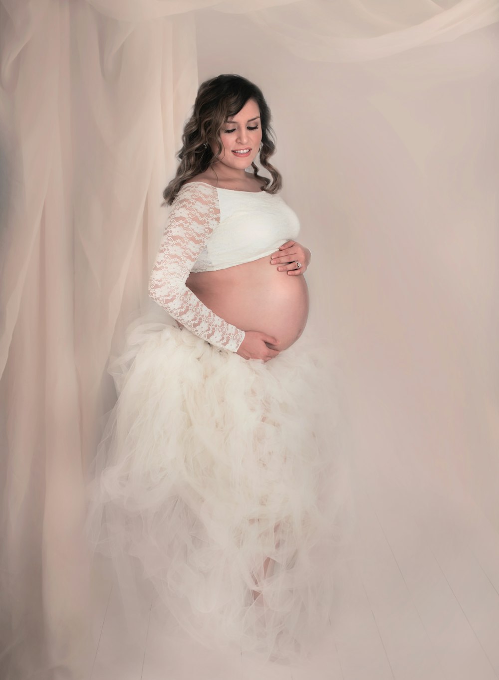 femme enceinte portant un crop top blanc à manches longues et une jupe blanche à volants debout contre un mur recouvert de blanc