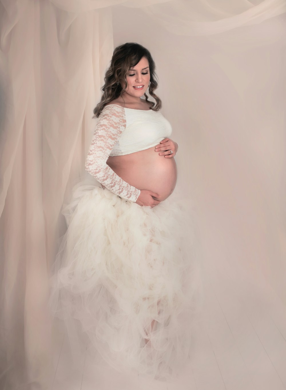 pregnant woman wearing white long-sleeved crop-top and white ruffled skirt standing against white covered wall