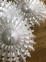 White paper decorations resembling snowflakes are placed on a wooden surface. The intricate patterns of the paper cutouts create a delicate and festive appearance.
