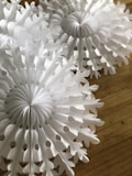 White paper decorations resembling snowflakes are placed on a wooden surface. The intricate patterns of the paper cutouts create a delicate and festive appearance.