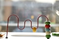A wooden toy with colorful beads threaded on a winding red metal wire. The beads are arranged in a vertical stack with a few separated along the wire path. The setup is placed on a smooth, reflective surface with a blurred background giving a sense of a bright, sunlit room.