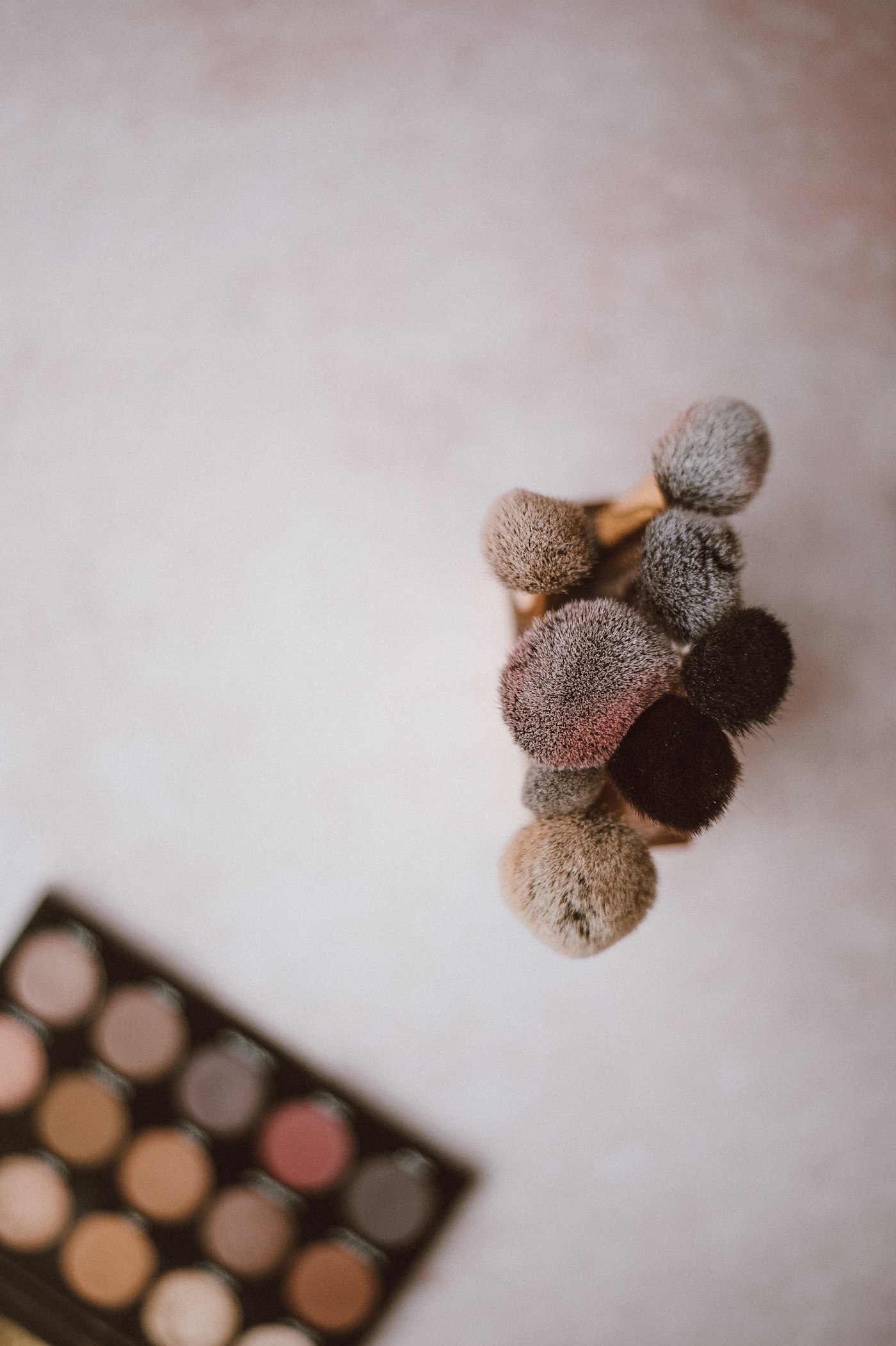 A close-up of several soft makeup brushes in neutral and muted shades on a pale surface, with part of a colour palette visible in the lower-left corner.