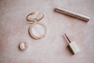 A flat lay arrangement of makeup items including an open compact with multiple shades of powder, a small pot of glitter, a tube labeled mascara, and a lip gloss wand. The items are placed on a beige textured surface.