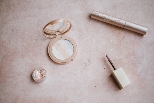 A flat lay arrangement of makeup items including an open compact with multiple shades of powder, a small pot of glitter, a tube labeled mascara, and a lip gloss wand. The items are placed on a beige textured surface.