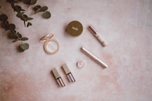 Various cosmetic items are arranged on a light, textured surface alongside fresh green foliage. The products include a circular compact with a mirror, a concealer, a small jar of glitter, a mascara, and two liquid products with shiny caps. The arrangement is minimalistic and aesthetically pleasing.