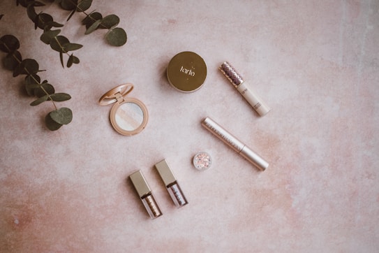 Various cosmetic items are arranged on a light, textured surface alongside fresh green foliage. The products include a circular compact with a mirror, a concealer, a small jar of glitter, a mascara, and two liquid products with shiny caps. The arrangement is minimalistic and aesthetically pleasing.