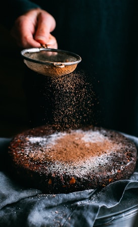 A chocolate cake is being dusted with cocoa powder using a sieve held by a hand. The cake sits on a cloth-covered surface, and the light casts a warm, inviting glow over the scene, highlighting the textures of the cake and the falling cocoa powder.