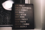 A motivational sign with various inspirational phrases such as 'LIVE FULLY', 'CREATE HAPPINESS', and 'LOVE DEEPLY' is placed next to a window with raindrops visible on the glass. The setting is dimly lit, creating a cozy and reflective atmosphere.